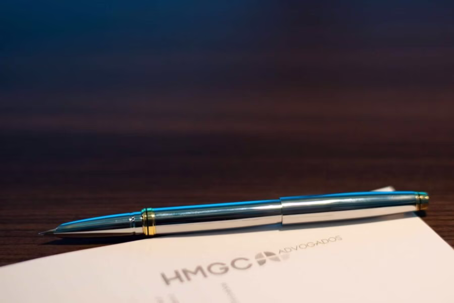 Caneta azul e dourada sobre documento com logo HMGC Advogados em superfície de madeira escura.